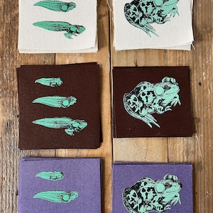 May include: Square fabric patches with frog and tadpole designs. The patches are in white, brown, and purple. The frog and tadpole illustrations are in a light green color. The frog patches show a detailed side view of the frog.