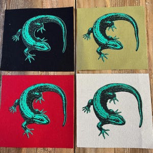 May include: Four square patches with a teal lizard design. The patches are on black, olive, red, and off-white backgrounds. The lizard is depicted with its tail curved over its body. The patches are likely for sewing or ironing onto clothing or other fabric items.