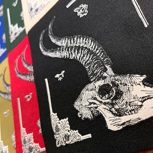 May include: A collection of square fabric patches featuring a white ink illustration of a horned animal skull. The patches are available in various solid colors including black, red, gold, blue, green, orange, and purple, each with ornate corner details.