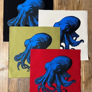May include: A collection of fabric squares featuring a blue octopus design. The squares are in various colors, including black, white, olive green, and red. The octopus illustration is detailed, with visible tentacles and a textured appearance. The fabric squares are arranged on a wooden surface.
