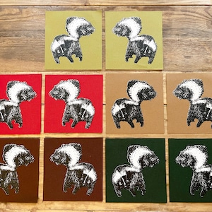 Könnte beinhalten: Eine Sammlung quadratischer Karten mit einer schwarz-weißen Stinktierillustration. Die Karten sind in Reihen und Spalten angeordnet, mit Hintergründen in verschiedenen Farben, darunter Rot, Grün, Braun und Beige. Das Stinktier-Design ist auf allen Karten einheitlich.