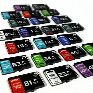 May include: A collection of black rectangular Formula 1 racing team nameplates, each with a driver's name, number, and team logo. The nameplates feature various color schemes and are arranged in a grid pattern.