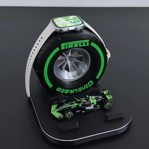 May include: A black and green tire-shaped Apple Watch charging stand with a miniature race car. The stand features the Pirelli and Cinturato logos in green. A silver Apple Watch with a white band rests on top of the tire.