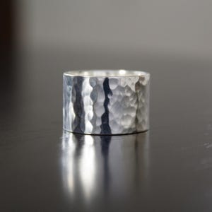 May include: A wide, silver ring with a hammered texture. The ring has a cylindrical shape and reflects light, creating highlights and shadows. The surface has a textured, irregular pattern, giving it a rustic appearance.