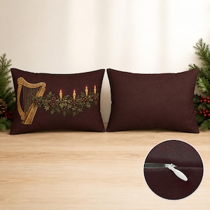 May include: Two rectangular dark brown throw pillows. The left pillow features a gold harp, green holly leaves, red berries, and four lit candles. The right pillow is solid dark brown. A circular inset shows a close-up of a white zipper closure.
