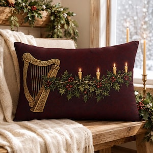 May include: A rectangular throw pillow in a deep burgundy color featuring a printed design of a gold Celtic harp on the left side and a horizontal garland of green holly leaves with red berries and four lit candles extending to the right.