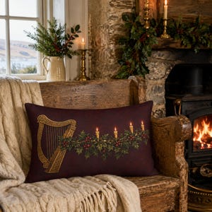 May include: A rectangular throw pillow in a deep burgundy color featuring a printed graphic of a golden Celtic harp and a festive garland of green holly leaves, red berries, and four lit taper candles arranged horizontally across the center.