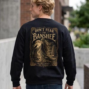 May include: A black crewneck sweatshirt featuring a gold-toned graphic on the back. The design depicts a banshee, a raven, and a full moon within an ornate frame, with the text "DON'T FEAR THE BANSHEE" and "SHE ONLY BRINGS THE MESSAGE" printed.