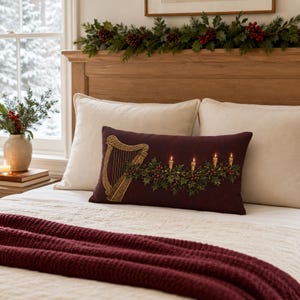 May include: A rectangular lumbar pillow in deep burgundy featuring a gold harp illustration and a printed design of green holly branches with red berries and four lit candles. The pillow is placed on a bed with cream pillows and a burgundy throw.