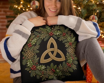 May include: A square throw pillow with a black background featuring a central gold triquetra knot symbol surrounded by a festive wreath of green pine needles, holly leaves, red berries, and small gold Celtic knot accents in the corners.
