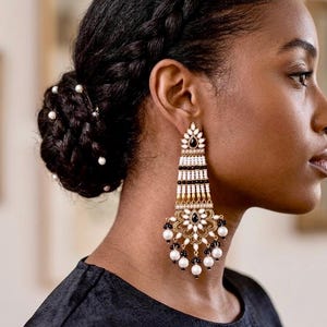 May include: Elegant chandelier earrings with a gold, black, white, and pearl design. The earrings feature a floral motif and dangling pearls. The earrings are long and ornate, perfect for special occasions.