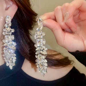 Op de afbeelding: Elegante oorbellen van heldere kristallen. De oorbellen hebben een cascade van kristallen in verschillende vormen, waaronder rechthoekige, ovale en bladvormige stenen. De kristallen zijn gezet in een zilverkleurig metaal, wat een sprankelende, glamoureuze uitstraling creëert.