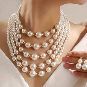 May include: A multi-strand pearl necklace with gold accents and matching pearl earrings. The necklace features graduated sizes of white pearls. The earrings have two white pearls each, connected by gold. The jewelry is displayed on a person wearing a white top.