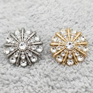 May include: Two decorative buttons with a floral design. One button is silver-toned, the other is gold-toned. Both buttons feature a central, large, clear rhinestone surrounded by smaller rhinestones.