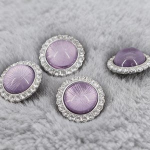 Peut inclure: Quatre boutons décoratifs avec un centre violet clair et une bordure argentée ornée de strass. Les boutons sont de formes variées, rondes et ovales, sur un fond gris doux.