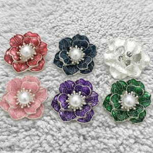 Puede incluir: Seis broches con forma de flor de varios colores, cada uno con un centro de perla. Los broches son de color rojo, rosa, azul, morado, blanco y verde. Cada uno tiene un diseño detallado de pétalos y un borde plateado.