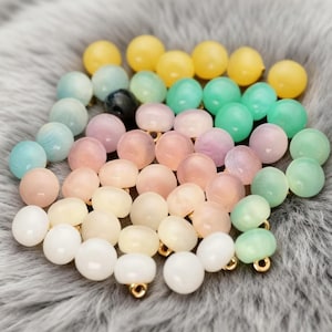 May include: A selection of pastel-coloured round beads in shades of yellow, green, pink, and blue. Each bead features a small gold-coloured loop. The beads are arranged on a soft, grey, textured surface.