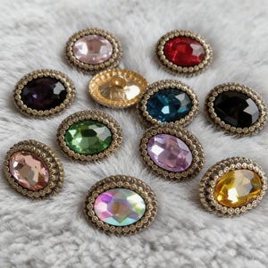 Könnte beinhalten: Eine Sammlung ovaler Edelstein-Knöpfe in verschiedenen Farben, darunter Rosa, Rot, Lila, Grün, Blau, Schwarz und Gelb. Jeder Knopf hat einen dekorativen, antikgoldfarbenen Metallrahmen. Die Knöpfe sind auf einer weichen, grauen Oberfläche angeordnet.