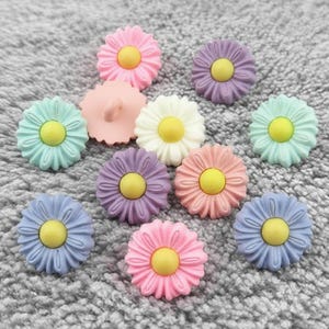 Può includere: Una collezione di bottoni a forma di margherita in varie tonalità pastello, tra cui rosa, viola, blu e verde menta, ciascuno con un centro giallo. Un bottone ha un gambo sul retro. I bottoni sono sparsi su una superficie grigia e strutturata.