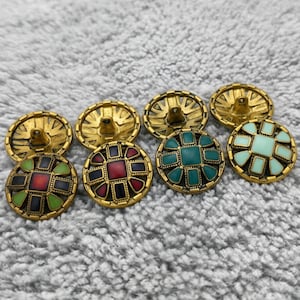 Può includere: Una collezione di bottoni decorativi con basi in metallo color oro. Le facce dei bottoni presentano disegni geometrici colorati in rosso, verde, blu e turchese. I bottoni sono rotondi e hanno un'estetica vintage.