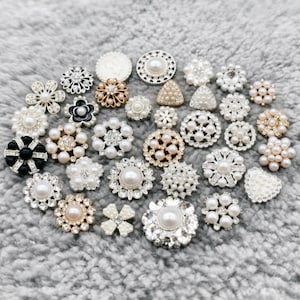 May include: A collection of decorative buttons in various shapes and sizes. The buttons feature pearls, rhinestones, and metallic accents in white, gold, and black. The buttons are arranged on a gray, textured surface.
