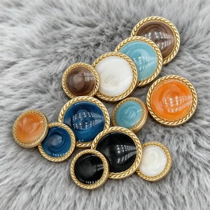 Puede incluir: Una colección de botones redondos en varios colores, incluyendo marrón, blanco, azul y naranja. Cada botón tiene un borde dorado en forma de cuerda. Los botones están dispuestos sobre una superficie suave y gris.