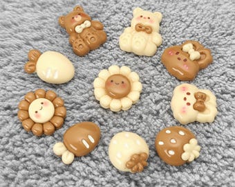 May include: A collection of small, decorative resin charms in various shapes and colors. The charms include teddy bears, flowers, and food items like strawberries and cookies. The color palette consists of brown, cream, and white.