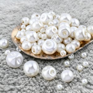 Può includere: Una collezione di perle bianche perlescenti di varie dimensioni, alcune con fori, è esposta in una conchiglia dorata. Le perle sono sparse su una superficie grigia e morbida, creando un contrasto strutturato.