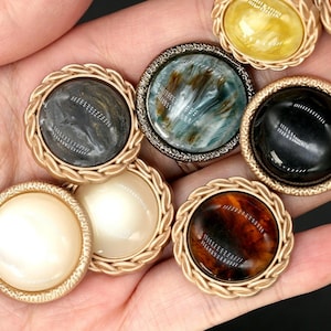 May include: A collection of round buttons with various colors and textures. The buttons have a gold-colored rope-like border. Colors include black, cream, brown, and blue-green. The buttons are likely for sewing or crafting projects.