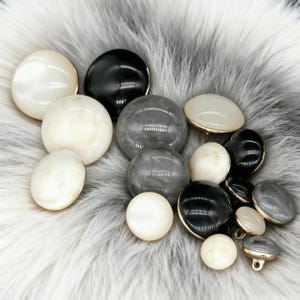 May include: A selection of buttons in white, black, and grey. Some buttons have a gold rim. The buttons are round, oval, and spherical, with a glossy finish. The buttons are scattered on a fluffy, white and grey surface.