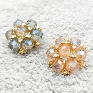 May include: Two circular decorative buttons featuring faceted crystal beads in shades of blue-grey and peach-gold. Each button is set in a gold-tone metal base with small clear rhinestones interspersed between the larger, multi-faceted glass beads.