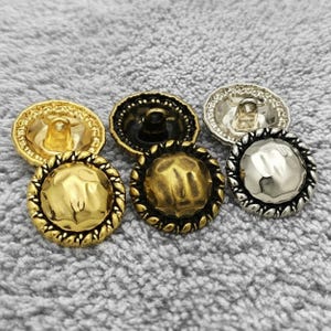May include: Close-up of six decorative buttons in various metallic finishes. The buttons feature gold, bronze, and silver tones with textured and rope-like edge designs. The buttons are for sewing or craft projects.