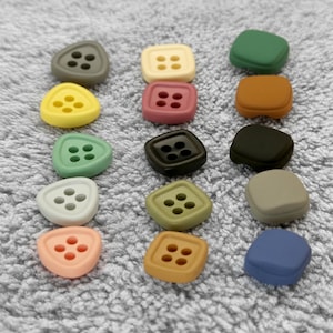 May include: A collection of colorful buttons in various shapes, including triangles and squares. The buttons are in shades of green, yellow, pink, and blue, with four holes each. The buttons are arranged on a textured, gray surface.