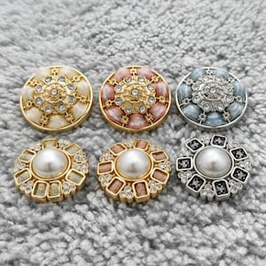 Può includere: Sei bottoni decorativi con disegni ornati. I bottoni presentano toni oro, rosa e blu, con accenti di perle e strass. I bottoni sono rotondi e hanno un motivo floreale dettagliato.