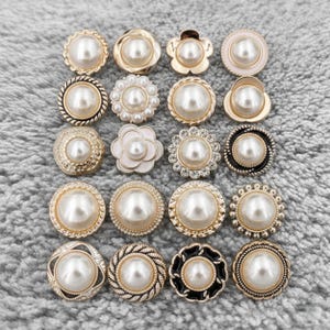 May include: A selection of decorative buttons featuring faux pearl centres and a variety of gold, black, and white frames. The buttons come in diverse shapes, including round, floral, and scalloped designs. The buttons are displayed on a grey, textured surface.