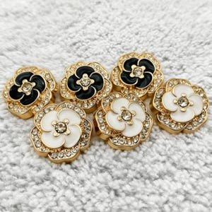 May include: A collection of six flower-shaped buttons featuring gold-tone metal edges, clear rhinestones, and enamel centers. Three buttons have black enamel petals, and three buttons have white enamel petals, each with a central clear rhinestone.