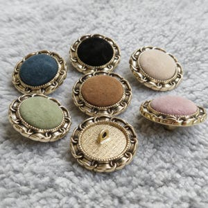May include: A selection of decorative buttons featuring gold-tone metal frames and fabric centers in various colours. The fabric inserts include black, brown, green, pink, and beige. The buttons have a vintage or antique appearance.