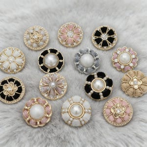May include: A selection of decorative buttons with floral designs. The buttons are in various colours, including gold, black, pink, white, and beige. Some have pearl or rhinestone accents, while others have a velvet or fabric texture.