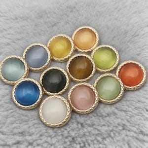 Op de afbeelding: Een verzameling ronde knopen in verschillende kleuren, waaronder blauw, groen, geel en oranje. Elke knoop heeft een goudkleurige, getextureerde rand. De knopen zijn gerangschikt op een zacht, grijs oppervlak.