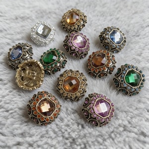 May include: A collection of decorative buttons with ornate metalwork and colorful faceted gemstones. The buttons feature various jewel tones, including blue, green, purple, and amber, set within detailed metal frames. One button has a lion's head design.