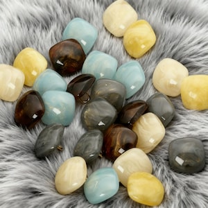 May include: A selection of square buttons in shades of light blue, yellow, cream, brown, and grey. The buttons have a glossy finish and are arranged on a soft, grey, furry surface. Some buttons feature a gold-coloured shank.