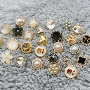 Könnte beinhalten: Eine Sammlung dekorativer Knöpfe in verschiedenen Formen und Designs. Die Knöpfe weisen Perlen, Kristalle und florale Motive mit Gold- und Silberakzenten auf. Einige Knöpfe haben eine Herz- oder Blumenform, andere sind rund oder quadratisch.