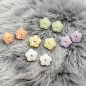 Op de afbeelding: Een verzameling bloemvormige kralen in verschillende pastelkleuren, waaronder perzik, groen, geel, wit en lavendel. Elke kraal heeft een klein goudkleurig midden. De kralen liggen op een zacht, grijs, harig oppervlak.