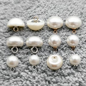 Puede incluir: Una colección de botones blancos con aspecto de perla, de varias formas y tamaños. Algunos botones tienen forma de cúpula, mientras que otros son esféricos. Cada botón tiene un ojal metálico para coser. Los botones están sobre una superficie gris texturizada.