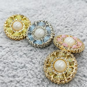May include: Four decorative buttons with a floral design. The buttons feature a central pearl surrounded by a ring of rhinestones and colorful enamel. Colors include yellow, blue, pink, and gold.