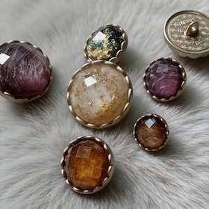 Op de afbeelding: Een verzameling decoratieve knopen met juweelachtige centra in tinten paars, goud en amber. De knopen hebben facetontwerpen en zijn gezet in sierlijke, goudkleurige metalen frames. Sommige hebben glitteraccenten.