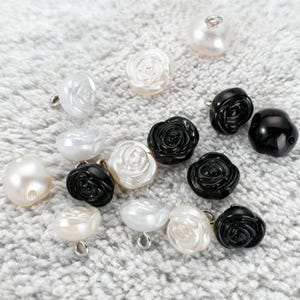 Puede incluir: Una colección de botones pequeños y redondos con forma de rosa en acabados negro brillante y blanco nacarado. Cada botón presenta un diseño de pétalos florales en relieve y una pequeña anilla metálica en la parte posterior para su fijación.