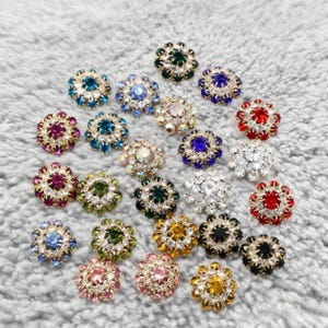 May include: A collection of decorative flower-shaped buttons with sparkling rhinestone centers and metallic edges. The buttons come in various colors, including blue, green, red, and gold. These embellishments are ideal for adding a touch of glamour to clothing or crafts.