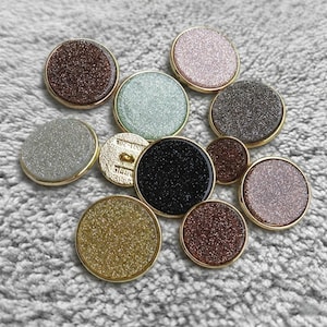 May include: A collection of round buttons in various colors, including brown, silver, black, and gold, with a glittery texture. Each button has a gold-colored metal rim. The buttons are arranged on a gray, textured surface.
