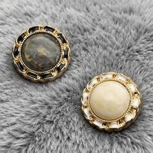 May include: Two decorative buttons with gold-coloured rims and ornate detailing. One button features a marbled grey and gold centre, while the other has a cream-coloured centre. The buttons are set against a grey, textured background.